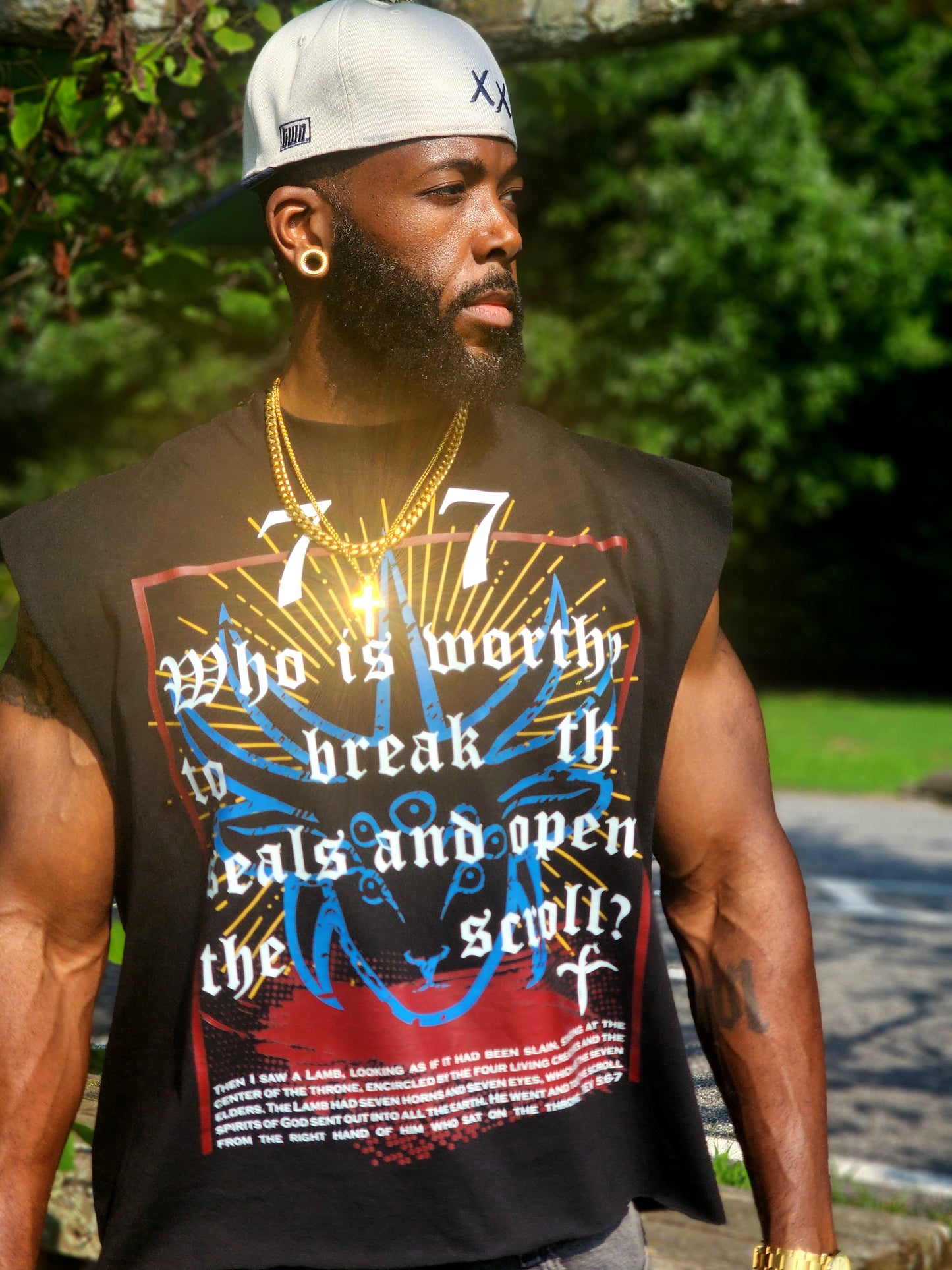 Model wearing Destroy This Temple "Revelation 5" TShirt, gray jeans, a white cap, and sunglasses, standing outdoors in front of a green background.