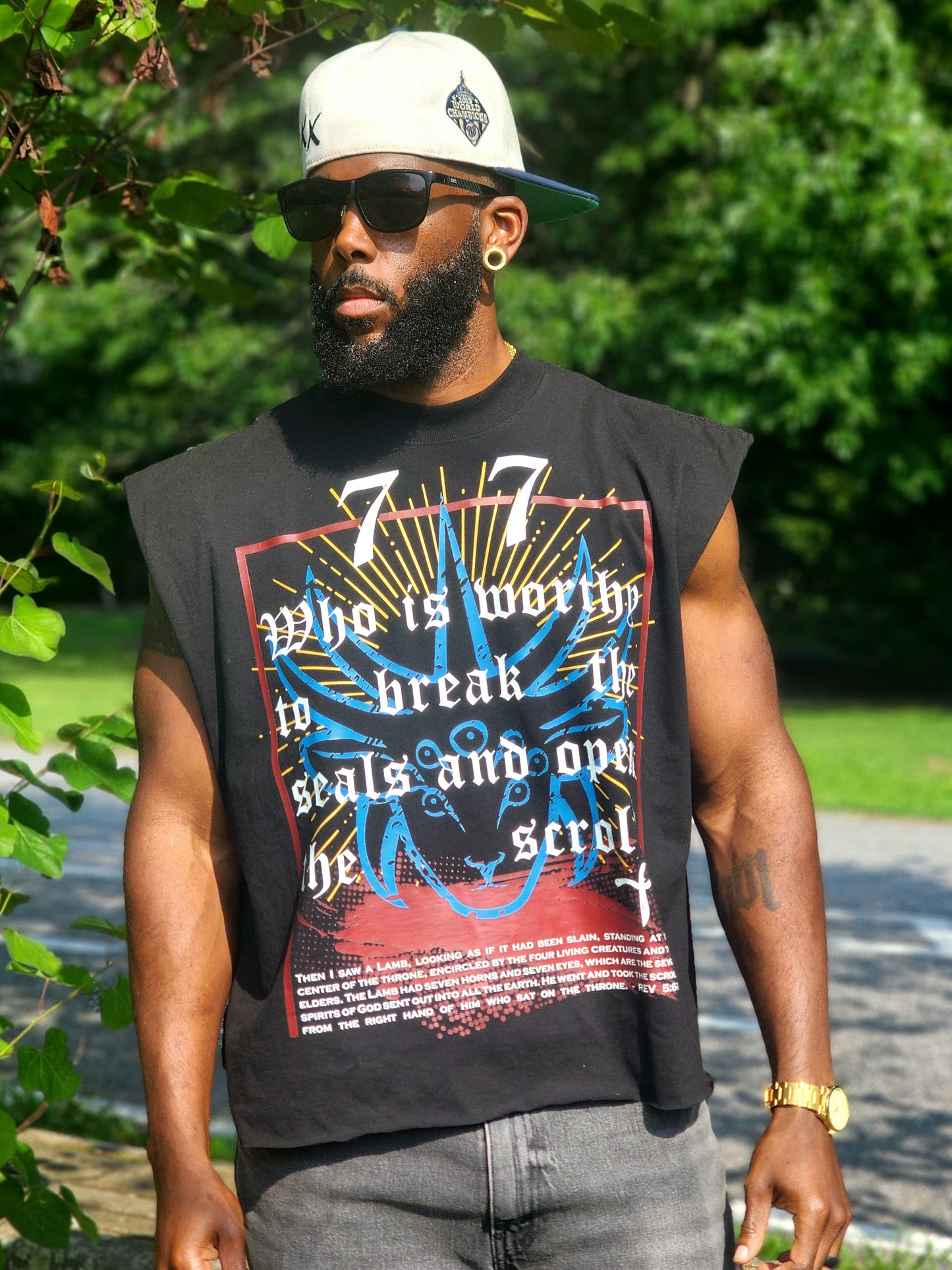 Model wearing Destroy This Temple "Revelation 5" TShirt, gray jeans, a white cap, and sunglasses, standing outdoors in front of a green background.