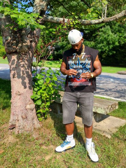 Model wearing Destroy This Temple "Revelation 5" TShirt, gray jeans, a white cap, and sunglasses, standing outdoors in front of a green background.